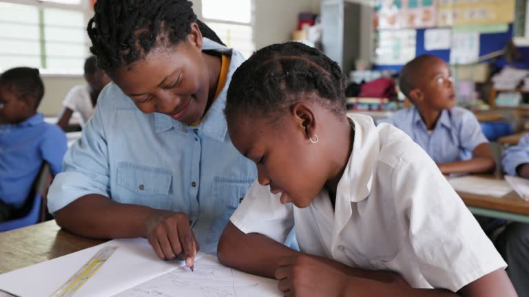 Close-up. Black African teacher sitting next to a young African girl at her desk in a classroom in Africa