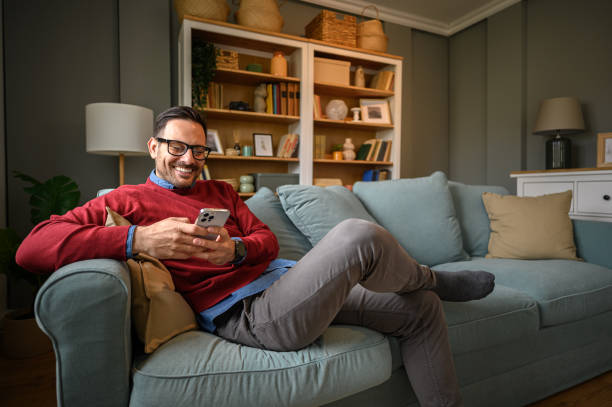Handsome freelancer smiling and doing social media marketing over mobile phone while sitting on sofa