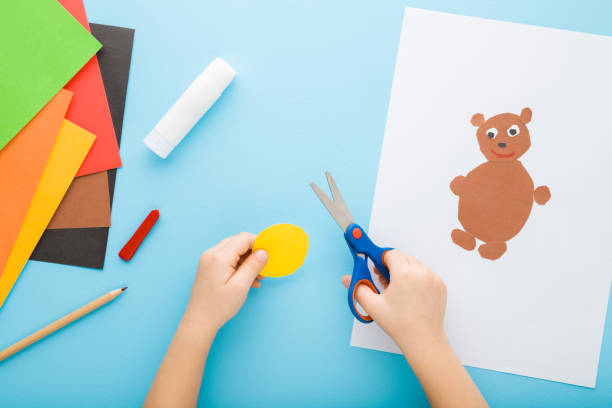 istock Child hands holding scissors, cutting application paper and creating teddy bear on white sheet. Blue table background. Pastel color. Point of view shot. Closeup. Toddler development. Top down view. 1692155643