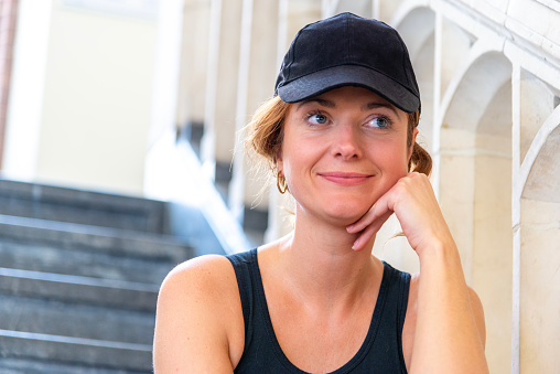 Portrait of a beautiful hipster woman with a black cap, situated on a staircase, looking at away. Smiling.