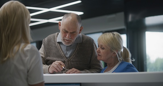 Elderly couple stand near reception desk in clinic lobby area, fill out papers. Female administrator talks to patients at information counter. Medical staff work in modern hospital or medical center.