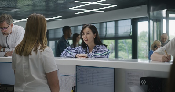Adult woman stands near reception desk in clinic lobby area and comes to appointment with doctor. Female administrator talks with patient at information counter. Medical staff work in modern hospital.