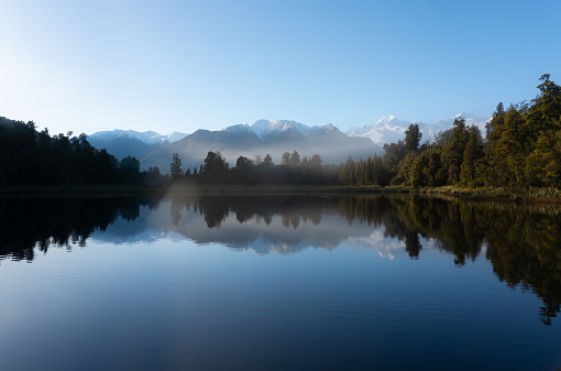Dawn breaks over spectacular Lake Matheson in New Zealand, whilst beyond we see the snow capped peaks of the Southern Alps.