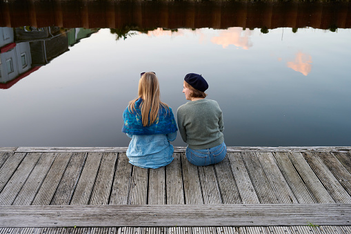 Two adult women having a nice time at the waterfront. Rear view portrait of women in conversation in front of water reflecting sky