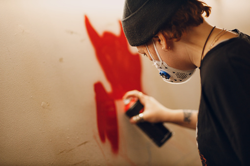 Young woman with a spray balloon with paint in her hand in a protective mask painting wall garage street art graffiti