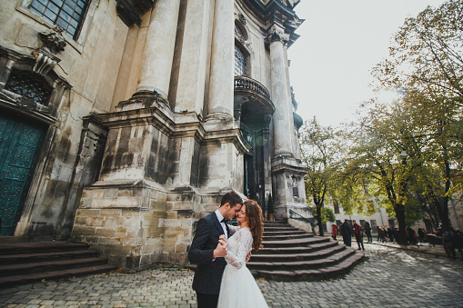 Wedding couple hugs in the old city. Stone walls of ancient church. Rustic lace bride with hair down and groom in grey suit and bow tie. Romantic love in vintage atmosphere street. Gothic cathedral.