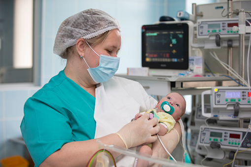 Neonatal Intensive Care Unit. The doctor holds a newborn child in his arms against the background of medical devices.