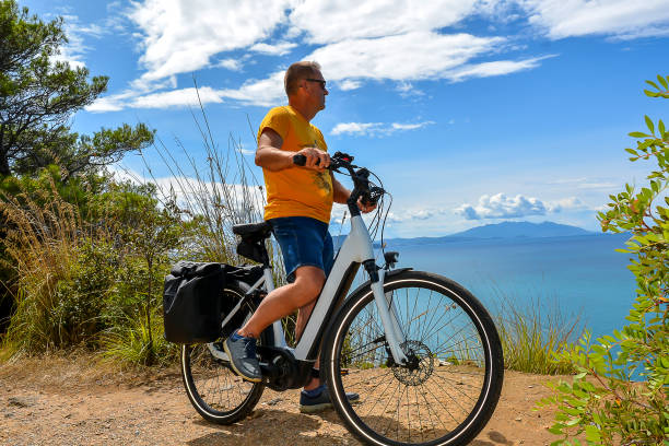 A middle-aged man wearing denim shorts and a yellow T-shirt on an electric bike stops and admires the beautiful scenery on the Italian coast in Calamaresca Bay, Tuscany, during his bike tour. stock photo