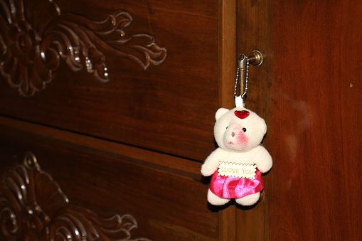 A teddy is hanging on a wooden door handle with carved design background, the stuffed bear is looking nice with the wooden cupboard which is hanging on the door, the handmade bear is looking very clear and bright in the room.