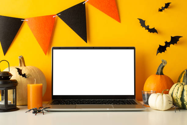 istock Spooky office side view setup: Laptop on table surrounded by Halloween decor - pumpkins, bats, spiders, candles, and lamp, against yellow wall with a festive flag garland. Empty screen for advert 1692386374