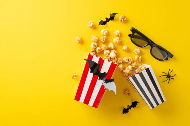 istock Halloween cinematic delight with friends. Top view angle captures themed decor: popcorn boxes filled with treats, creepy insect, flying bats, and 3D glasses. Yellow setting boasts space for text or ad 1692386488