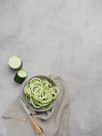 Raw spiralized zucchini noodles in a bowl, next to knife on a gray cement table with copy space