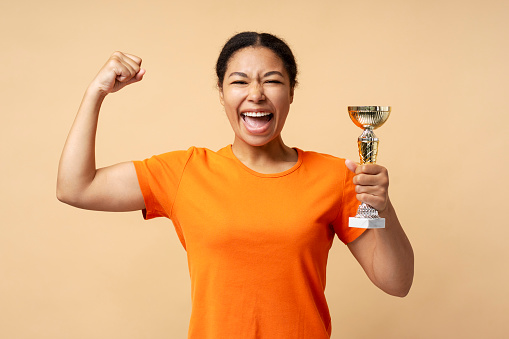 Young overjoyed African American woman holding trophy cup, celebration success isolated on beige background. Sport victory, winning competition concept