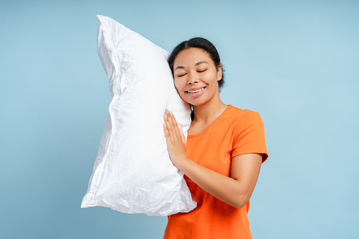 Happy African American woman sleeping, holding white pillow isolated on blue background. Morning concept. Stock photo