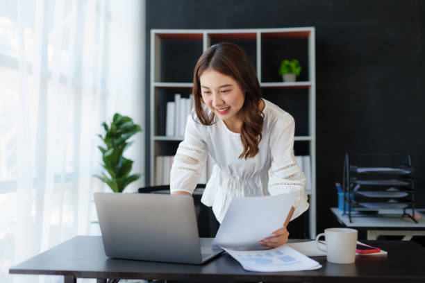 istock Beautiful young Asian businesswoman is saving and checking work and documents with her laptop computer at her desk with a bright smile on her face. 1692400394