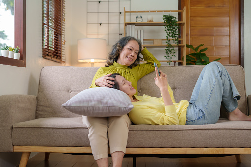Happy Asian mother and daughter taking a selfie in the living room at home on the sofa on vacation with a bright smile. Happy Asian mother and daughter using smartphone at home.