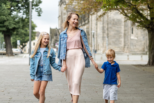 Young adult beautiful mom enjoy having fun walking with two kids in european city street at summer day. Happy parenthood family concept. Mother care resonsibility. People go outside in Germany.