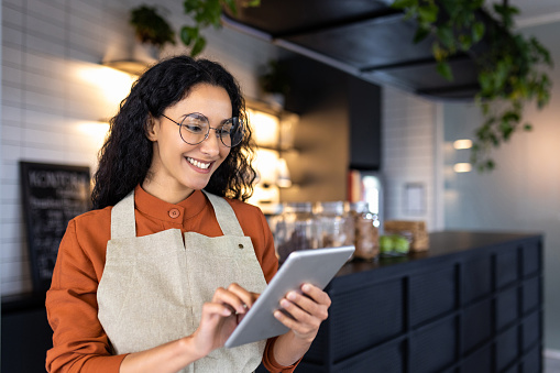 Female barista waitress inside cafe using tablet, latin american restaurant owner smiling using tablet app to confirm online order.