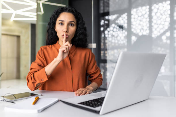 porträt einer lateinamerikanischen jungen geschäftsfrau, die im büro an einem laptop arbeitet und der kamera eine geste des schweigens zeigt. er schließt den mund mit dem finger - silence finger on lips businesswoman one person stock-fotos und bilder