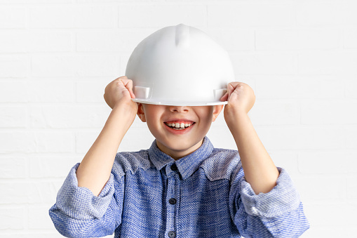 Laughing little boy holds a hardhat helmet with his hands covering his eyes. Emotional child laughs with teeth. Fun, dreams, childhood, imagination, renovation, construction, insurance concept.