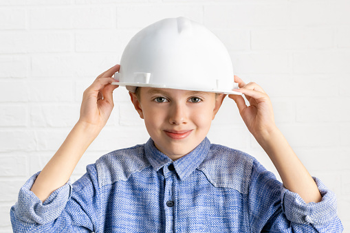Smiling boy holding a construction helmet with his hands on his head. Child pretends to be an engineer, architect or builder worker. Choice of profession, education and dreams of the future concept.