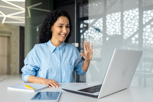 Young successful hispanic woman working inside office with laptop, waving at camera using video call, businesswoman talking with colleagues online remotely.