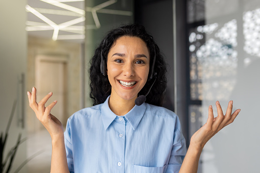 Webcam view, business woman looking at camera with headset talking to customers on video call, Hispanic woman inside office at workplace, advising consumers buyers online support service.