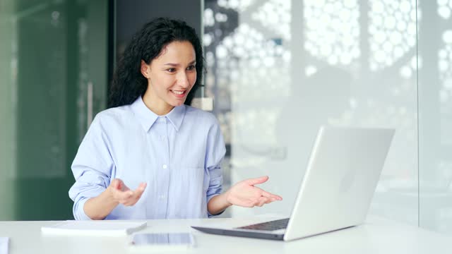 Confident young female employee talking on a video call using a laptop sitting at a workplace in a modern office