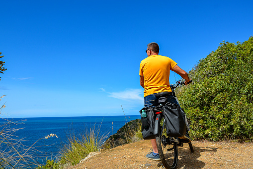 A middle-aged man wearing denim shorts and a yellow T-shirt on an electric bike stops and admires the beautiful scenery on the Italian coast in Calamaresca Bay, Tuscany, during his bike tour.