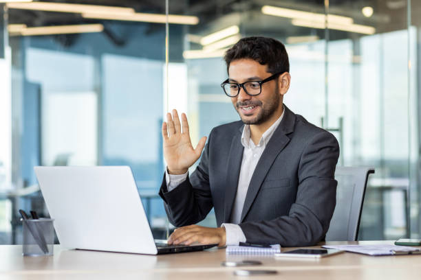 istock Young arab businessman inside office workplace using laptop for remote communication video call, man greeting and smiling, waving hand gesture of greeting and friendship 1692546062