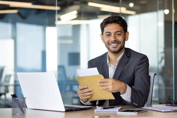 istock Portrait of young successful businessman at workplace, man received mail envelope notification letter, man smiling and looking at camera, satisfied with achievement results boss in business suit 1692546081