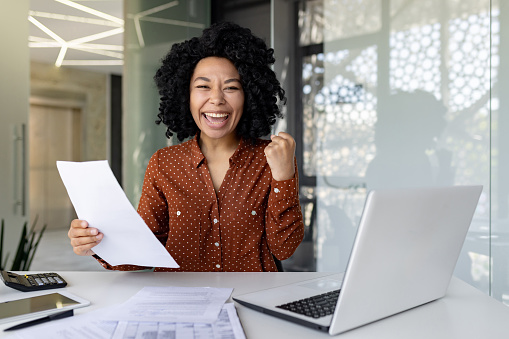 Portrait of successful african american woman boss at workplace, businesswoman behind paper work smiling and looking at camera, holding hand up winner and triumphant gesture inside office