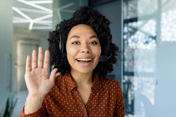 istock Video call online remote chat, businesswoman inside office smiling and looking at web camera with headset phone, waving hand gesture of greeting 1692577258