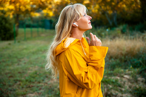 Blonde Woman with in ears headphones in yellow raincoat walking outdoors.