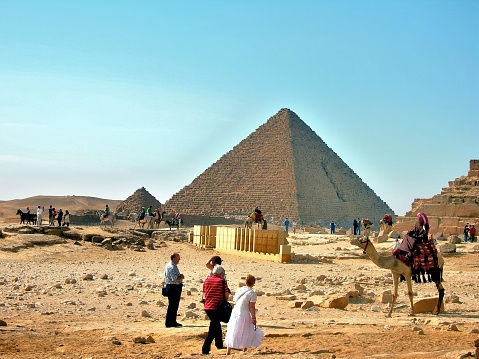 Giza,Egypt - December 2,2007 : A woman tourist in white clearly doesn’t have on the shoes for walking in the sand while a native man sits on his camel watching, perhaps getting a story to tell his friends riding the other camels and horses at the site