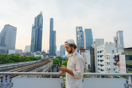 Young Caucasian man in cap  with smartphone  standing on the background of Bangkok cityscape