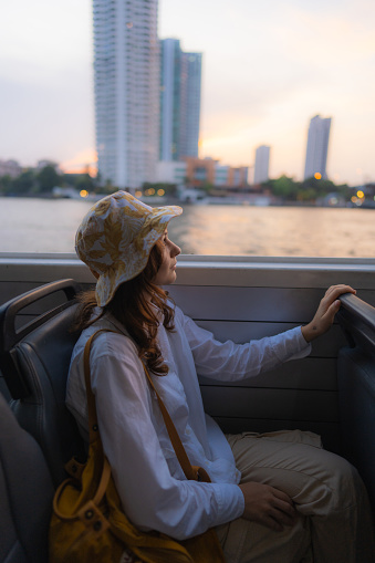 Young cheerful woman  traveling with boat on Chao Phraya river and looking at scenic view of Bangkok at sunset