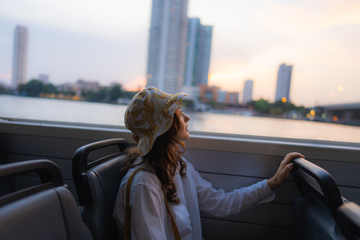 Young cheerful woman  traveling with boat on Chao Phraya river and looking at scenic view of Bangkok at sunset