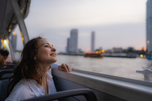 Young cheerful woman  traveling with boat on Chao Phraya river and looking at scenic view of Bangkok at sunset