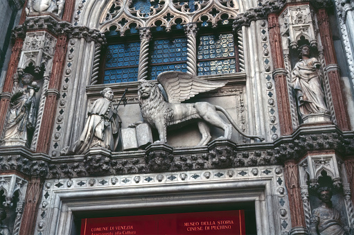 Venice, Italy- October 15, 1986: Close up of the statue of the winged lion of Saint Mark above the main entrance to the Doge's Palace; announcement for the Beijing Chinese History Museum exhibit.