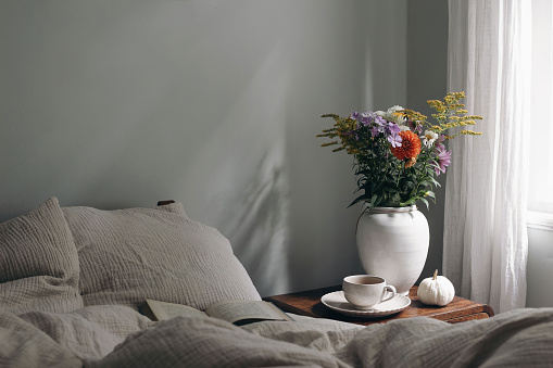 Elegant moody bedroom interior. Cup of coffee, pumpkin on retro wooden bedside table. White ceramic vase. Bouquet of dahlia, cosmos, solidago flowers, beige muslin cushions, book in bed near window.