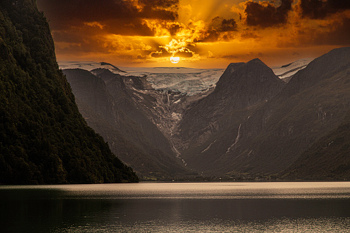 An enhanced shot of a Norwegian Fjord with a glacier at the of the Fjord. The sky has been made a deeper orange for effect. Three days of rain has swollen the rivers which run down the deep  sides of the mountains. Taken from the deck of cruise ship heading down the Fjord.