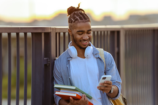 Smiling African America young male student holding books and using smart phone