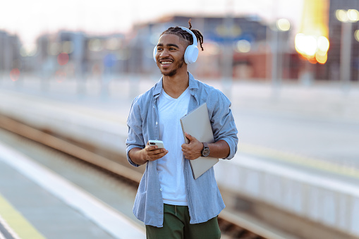 Smiling male student holding laptop and smart phone. He is waiting for a train on a train station at dusk