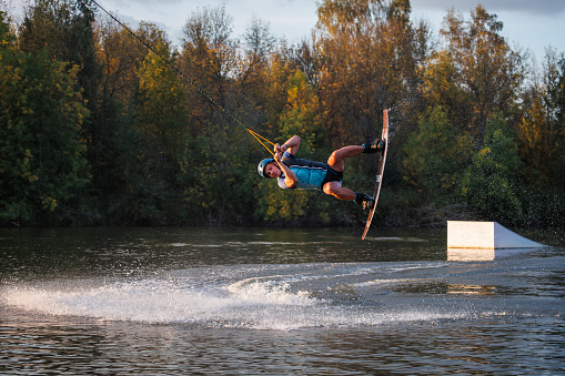 An athlete does a trick from a springboard. A rider jumps on a wakeboard against a background of a green forest. Sunset on the lake.