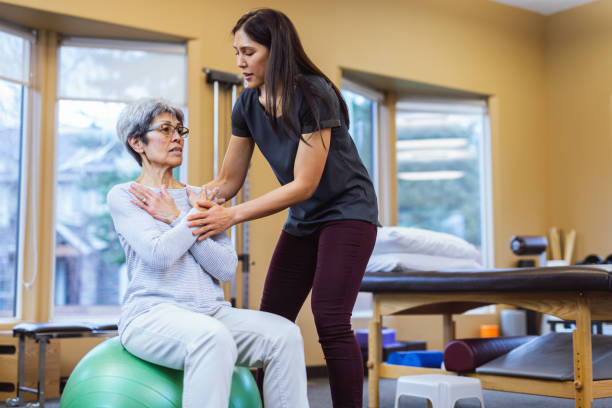 Senior patient sits on yoga ball with help of physical therapist stock photo
