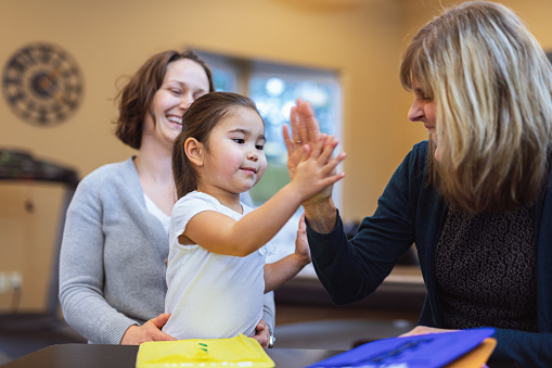Preschool age girl of Asian ethnicity high fives her proud, female physical therapist after completing a motor skills task in a health and wellness office.