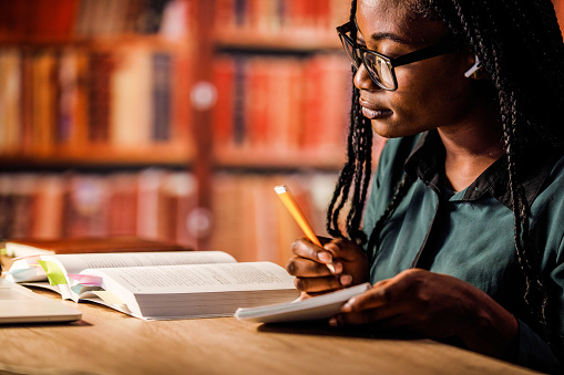 Copy space shot of diligent young woman sitting at table in a library, listening to music via blue-tooth headphones, reading a book and taking notes while studying for exam.