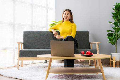 Beautiful happy smiling Asian woman relaxing using laptop computer in living room at home. Creative woman working online.