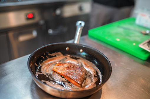 Close up shot of a salmon with a perfectly cooked crust. This sea food is inside a frying pan and is covered on a special sauce. There are cooking utensils around the kitchen counter.